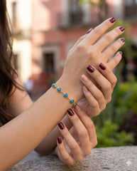 Manos cruzadas con pulsera ojo turco dorada, uñas vino, calle colonial | Gheller