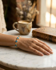 Pulsera ojo turco dorada sobre mesa de mármol con café y libro | Gheller Joyería